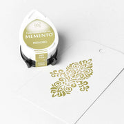 Ein pistaziengrünes Tsukineko Stempelkissen neben einem weißen Anhänger mit einem detailreichen Stempelmotiv in passender Tinte gestempelt.