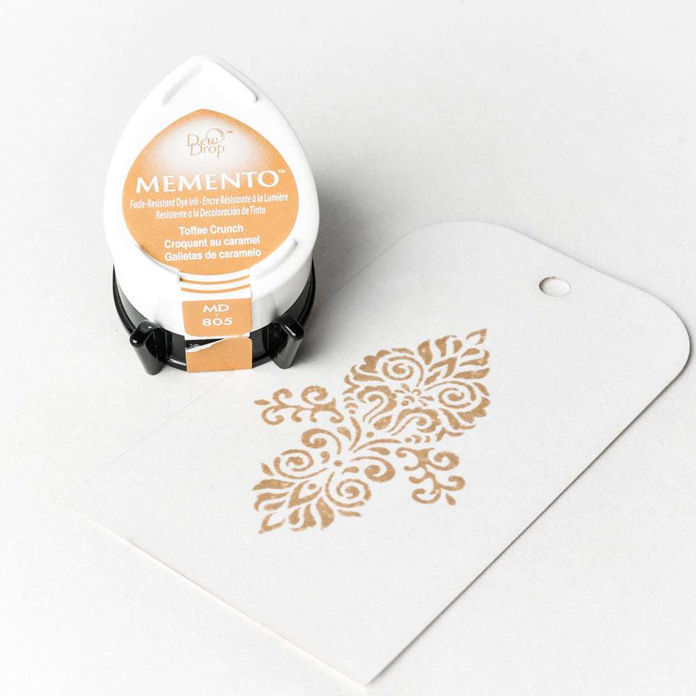 Ein Tsukineko Memento Stempelkissen und ein Anhänger mit einem verschnörkelten karamellfarbenen Stempelmotiv auf weißem Papier, hergestellt mit hochpigmentierter Farbe.