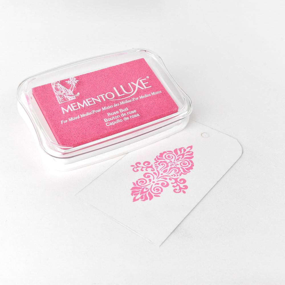 Ein rosafarbenes Tsukineko Stempelkissen und ein weißer Anhänger mit einem rosafarbenen, verschnörkelten Stempelmuster, perfekt für Pigment-Tinten-Projekte, auf weißem Hintergrund.