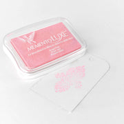 Ein rosafarbenes Tsukineko Memento Luxe Stempelkissen sitzt neben einem durchsichtigen Anhänger, der mit einem rosafarbenen Blumenmuster bestempelt ist. Perfekt, um Karten oder Textilien mit lebendigen Pigment-Tinten-Akzenten zu versehen.