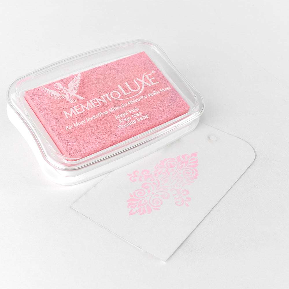 Ein rosafarbenes Tsukineko Memento Luxe Stempelkissen sitzt neben einem durchsichtigen Anhänger, der mit einem rosafarbenen Blumenmuster bestempelt ist. Perfekt, um Karten oder Textilien mit lebendigen Pigment-Tinten-Akzenten zu versehen.