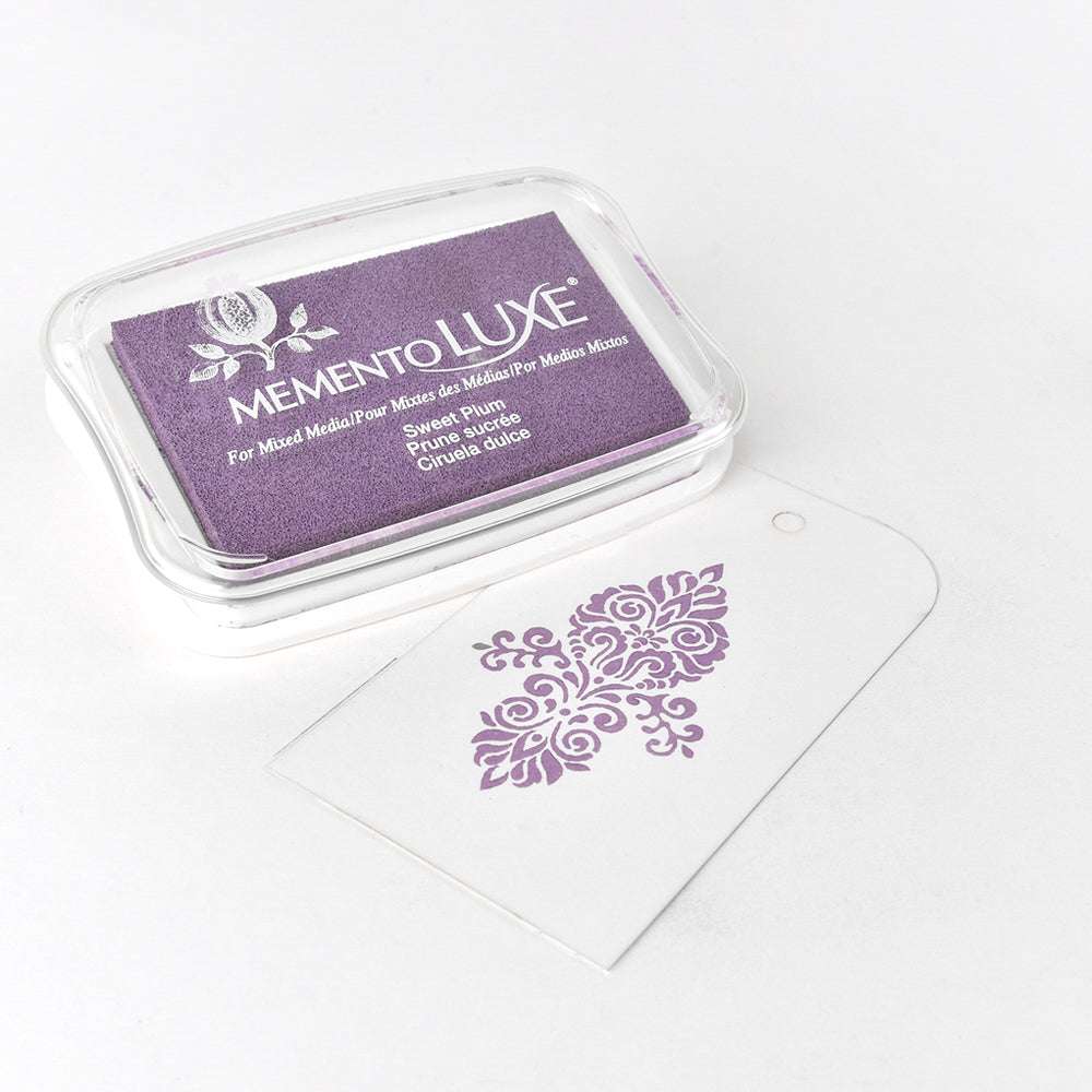Ein lilafarbenes Tsukineko Memento Luxe Stempelkissen liegt neben einem weißen Anhänger, der mit einem lilafarbenen Blumenmuster bestempelt ist. Die Pigment-Tinte ist vielseitig verwendbar und verleiht vielen Oberflächen ein satiniertes Finish.