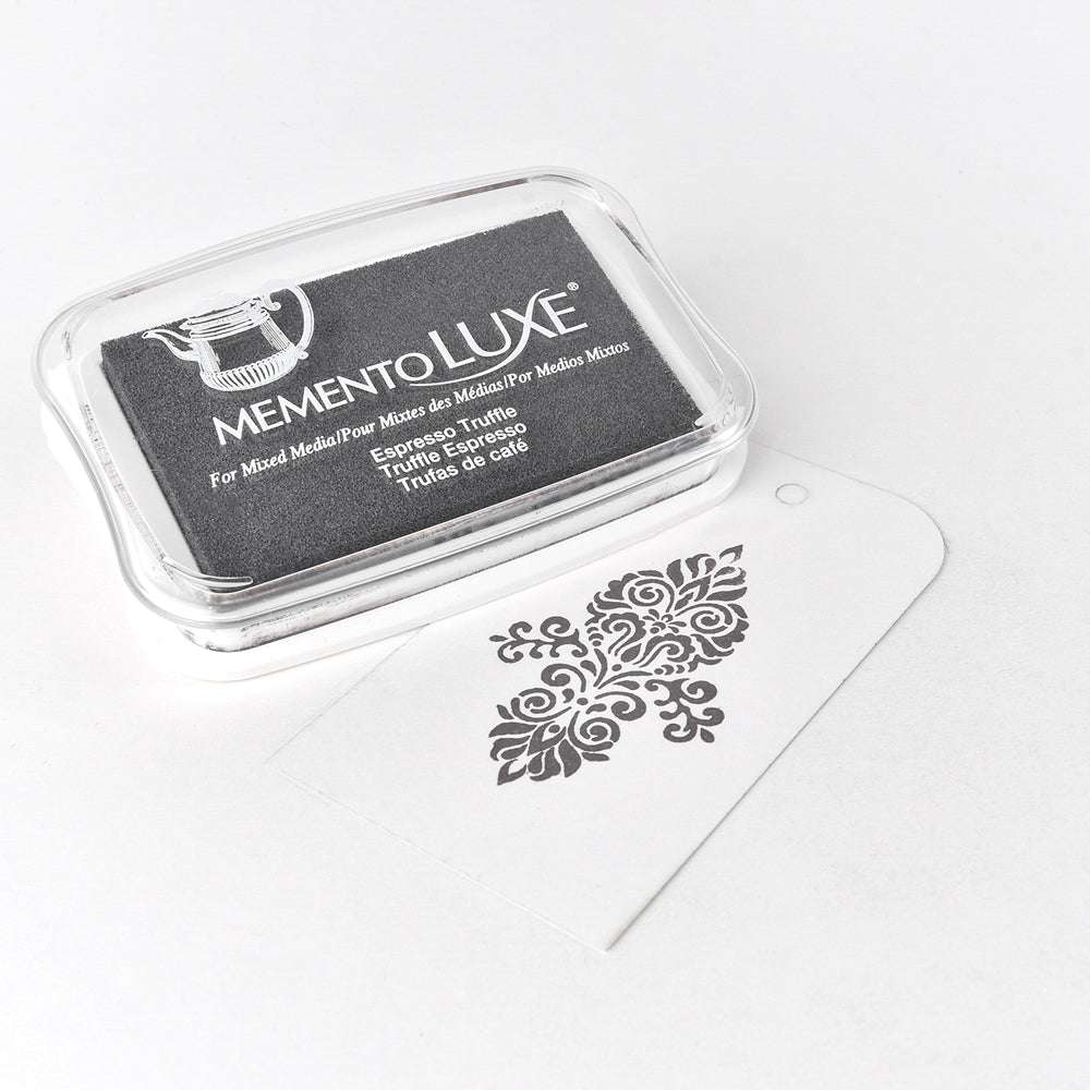 Das schwarze Pigmenttintenkissen Tsukineko Memento Luxe hat ein weißes Etikett mit einem verschnörkelten Blumenstempel und eignet sich perfekt für kreative Projekte und Textilstempelungen.
