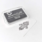 Ein schwarzes Tsukineko Memento Luxe Stempelkissen liegt neben einem weißen Anhänger mit dekorativem schwarzem Design - ideal zum Basteln mit satten Pigment-Tinten und zum Erkunden eines lebendigen Farbspektrums.