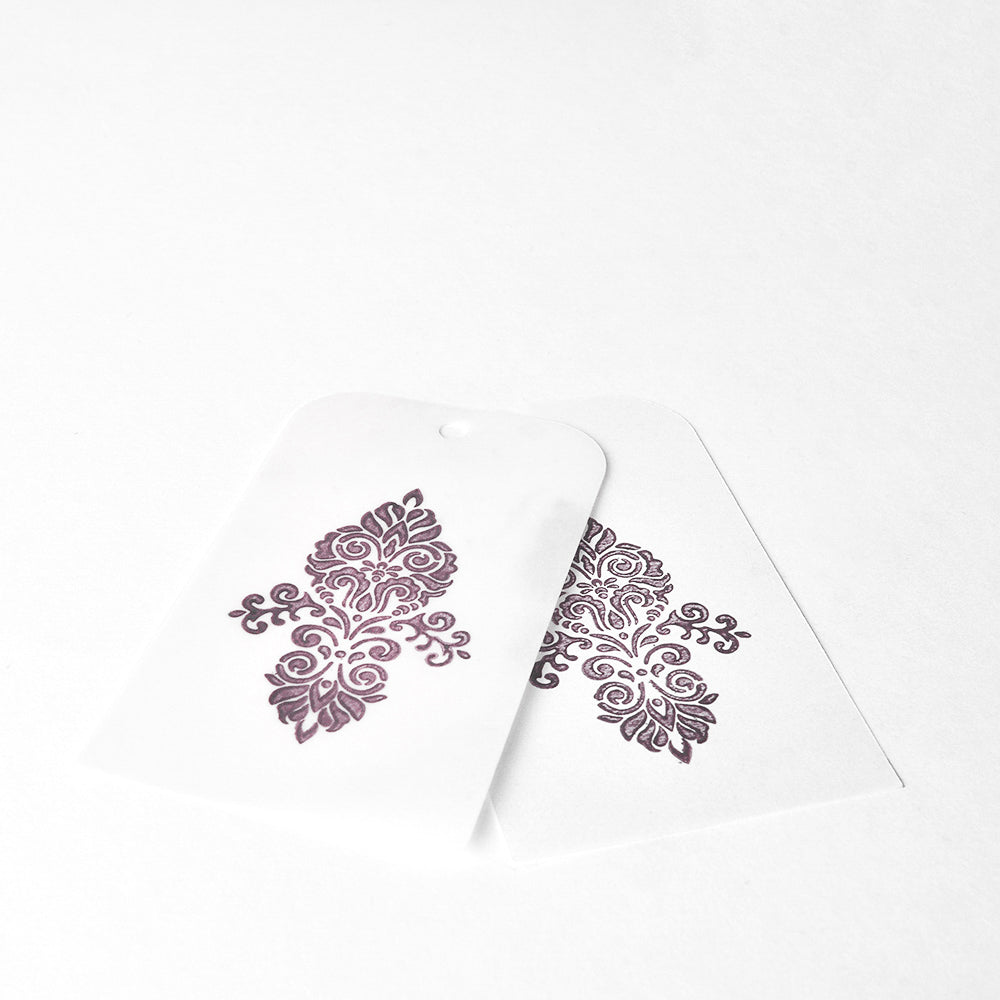 Zwei weiße Anhänger mit verschnörkelten dunkelvioletten Blumenmustern, die sich auf weißem Hintergrund überlappen, unterstreichen die reiche Wirkung der hochpigmentierten Tinte von Tsukineko Brilliance Stempelkissen.