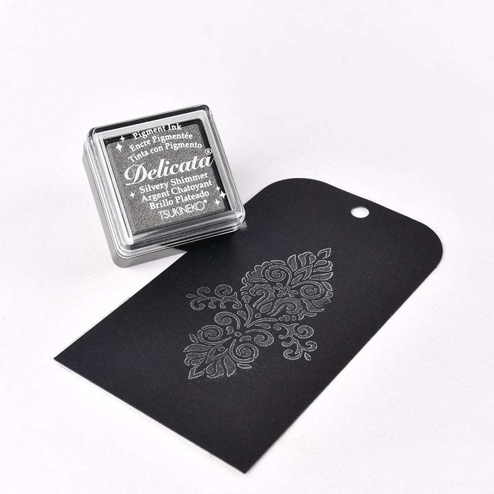 Ein schwarzer Geschenkanhänger mit silbernem Blumenmuster liegt neben einem Tsukineko Delicata Mini Stempelkissen, das sich perfekt eignet, um Ihren gestempelten Motiven metallische Effekte zu verleihen.
