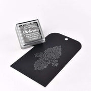 Ein Stempelkissen mit silberner Tinte von Tsukineko, ideal für metallische Effekte, liegt neben einem schwarzen Anhänger mit einem silbernen Blumenmuster.