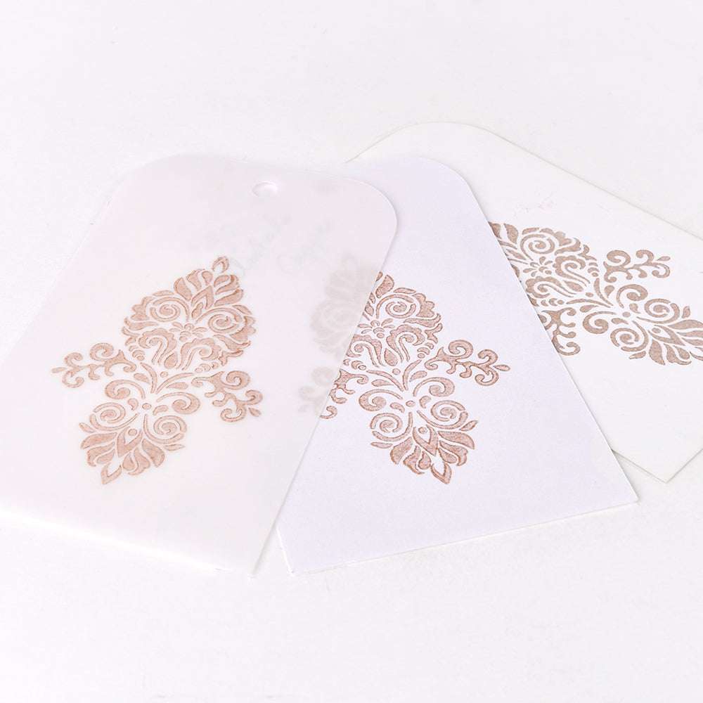 Drei Anhänger mit verschnörkelten goldenen Blumenmustern, erstellt mit Tsukineko Delicata Mini Stempelkissen für atemberaubende Metallic-Effekte - einer auf Transparentpapier und zwei auf weißem Cardstock, alle auf einer weißen Oberfläche.