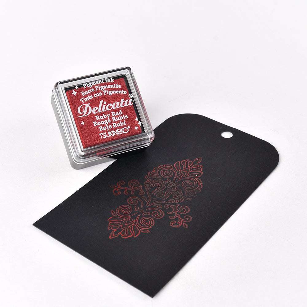 Tsukineko Red Delicata Mini Stempelkissen neben einem schwarzen Anhänger mit einem dekorativen roten Blumenmuster, das mit lichtechter Tinte und schimmernden metallischen Effekten bedruckt ist.