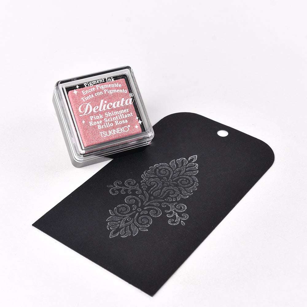 Das rosafarbene Tsukineko Delicata Mini Stempelkissen erzeugt elegante Metallic-Effekte mit einem gestempelten silbernen Blumenmuster auf einem schwarzen Anhänger - perfekt für Ihr Lieblings-Stempelmotiv.