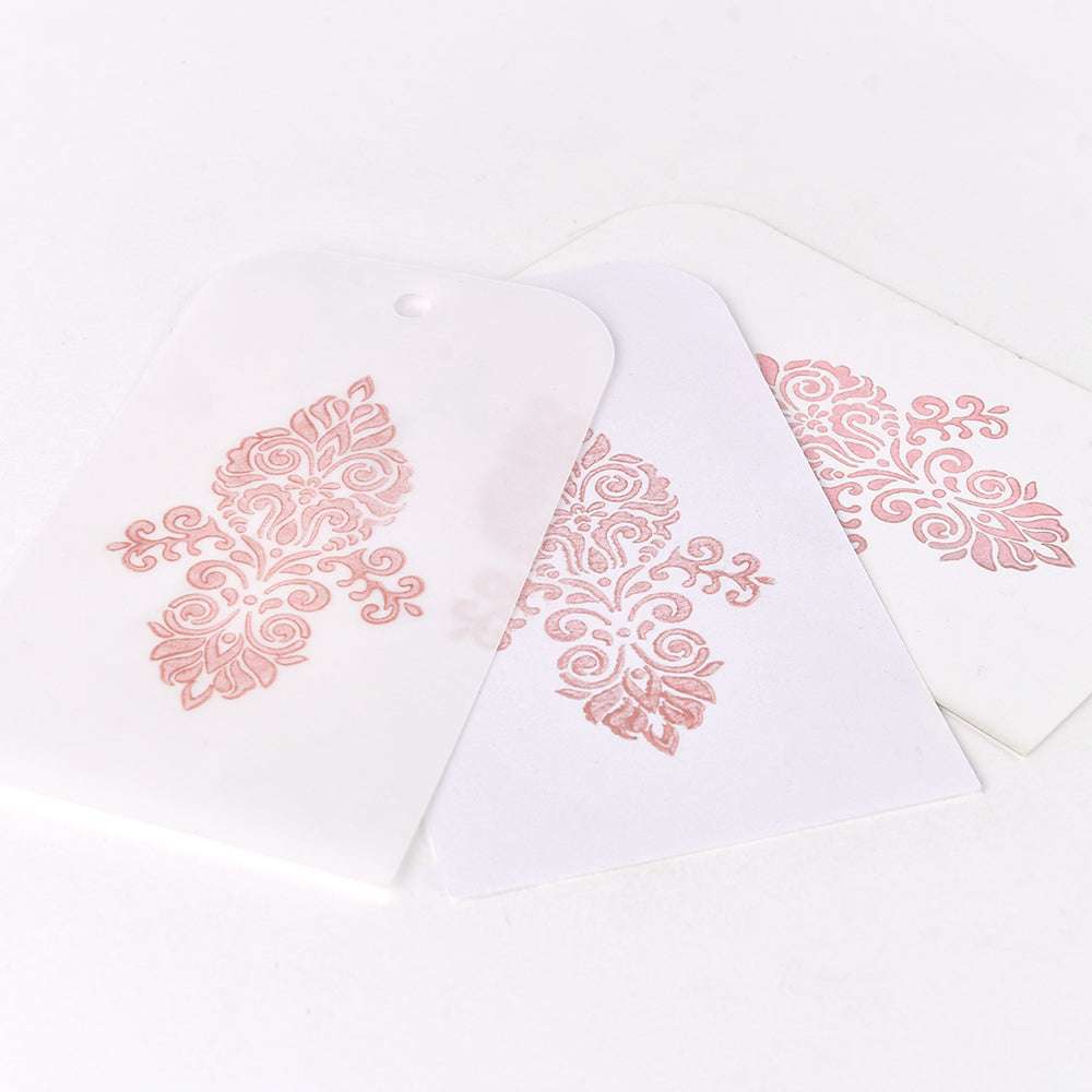 Drei weiße Anhänger von Tsukineko zeigen verschlungene rosafarbene Blumenmuster mit Metalleffekten, die überlappend auf einer weißen Fläche angeordnet sind.