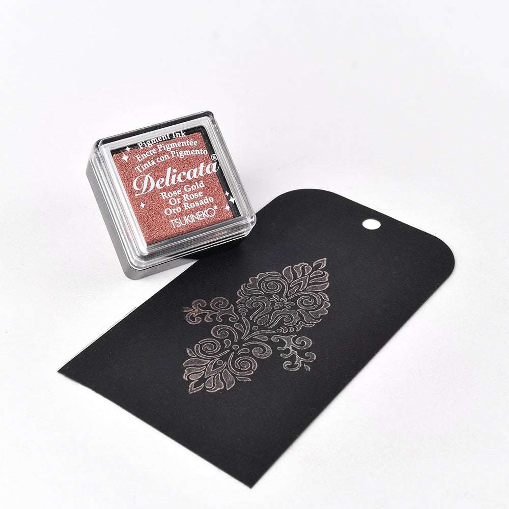 Ein roségoldenes Mini-Stempelkissen von Tsukineko liegt neben einem schwarzen Umschlag mit einem verschnörkelten Blumenmuster - ideal, um Ihren Basteleien metallische Effekte zu verleihen.