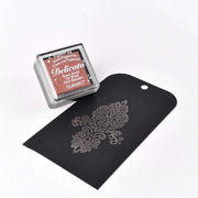 Ein roségoldenes Stempelkissen mit Metalleffekt von Tsukineko befindet sich neben einem schwarzen Umschlag mit einem verschnörkelten Blumenmuster.