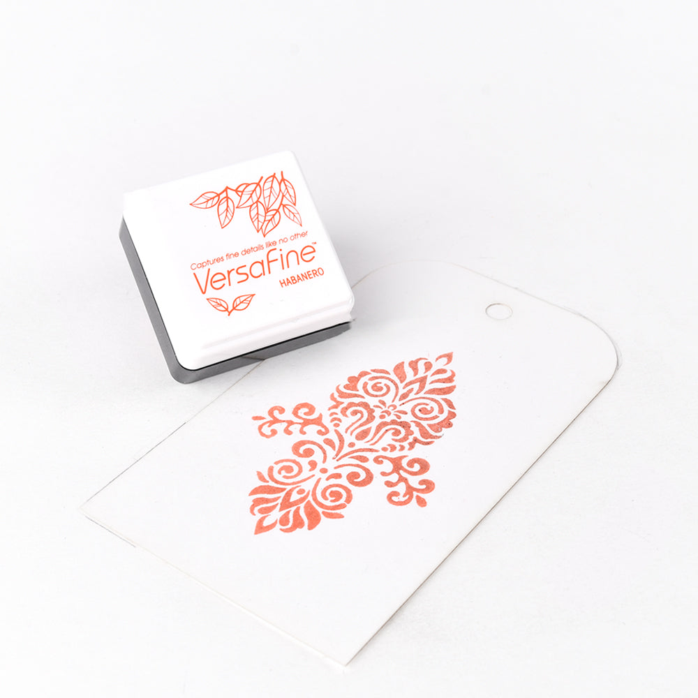 Ein kleines Tsukineko VersaFine Habanero Stempelkissen ist neben einem gestempelten, verschnörkelten Blumenmuster auf einem weißen Anhänger abgebildet. Mit diesem hochwertigen Versafine-Pigmenttintenkissen werden feine Details hervorgehoben.