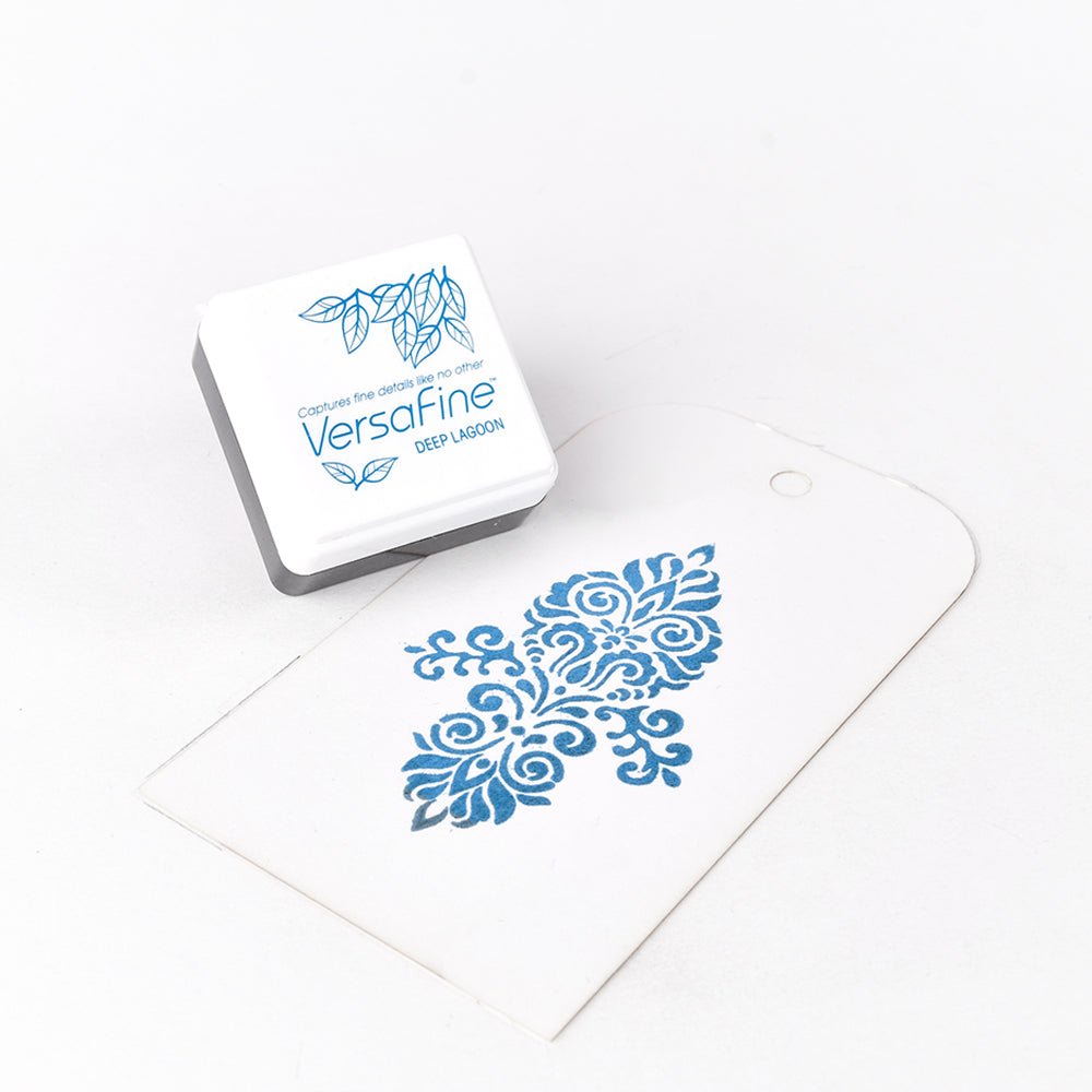 Ein Tsukineko Versafine Stempelkissen in Blau und ein weißer Tag mit feinem floralem Motiv durch Pigment-Tinte liegen auf weißem Untergrund. Perfekt zum Stempeln filigraner Details.