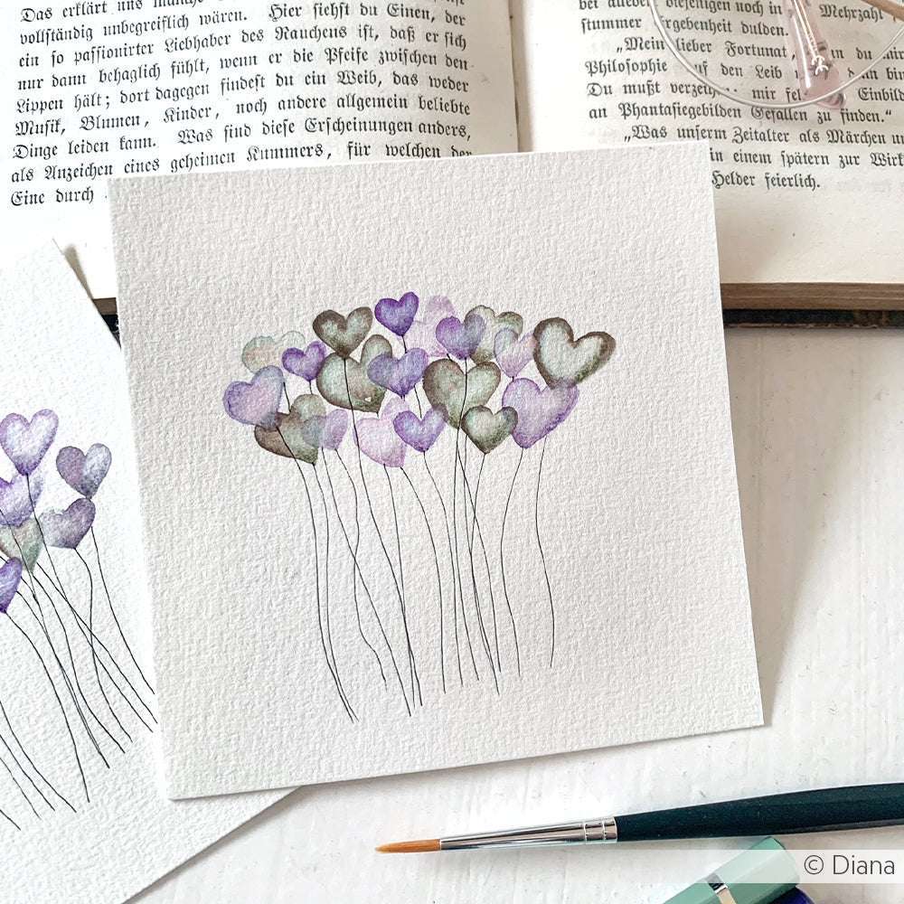 Neben einem aufgeschlagenen Buch, einem Pinsel und einem Kuretake Gansai Tambi Aquarellfarbe Farbkasten befindet sich ein Aquarellbild mit herzförmigen Blumen mit dünnen Stielen.