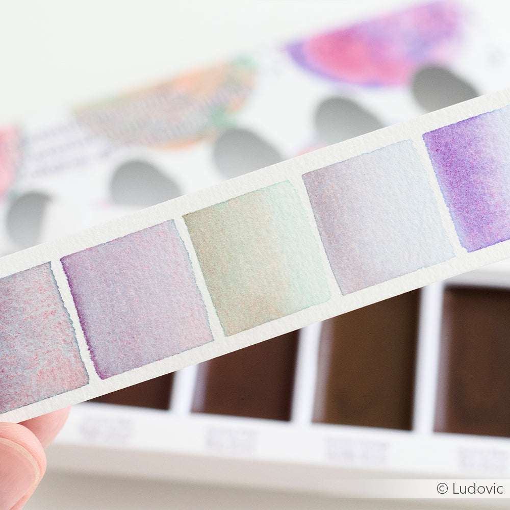 Eine Hand hält eine Musterkarte mit pastellfarbenen Aquarellquadraten; im Hintergrund erscheint ein verschwommenes Aquarellset von Kuretake Gansai Tambi.