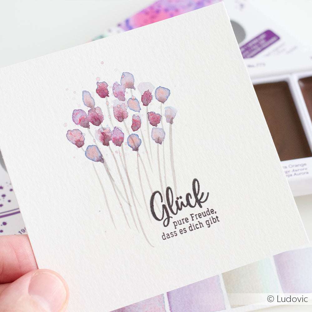 Eine Hand hält eine Karte mit Aquarellblumen, die mit der Aquarellfarbe Kuretake Gansai Tambi gemalt wurden, und mit dem deutschen Text: Glück, pure Freude, dass es dich gibt.