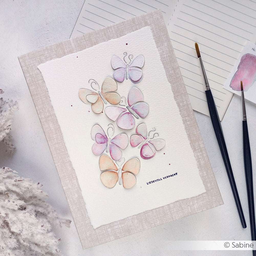 Aquarellmalerei von pastellfarbenen Schmetterlingen auf strukturiertem Papier, mit Pinseln und Notizpapier daneben. Mit zarten Effekten aus granulierter Wasserfarbe und leuchtenden Farbtönen aus Kuretake Gansai Tambi Aquarellfarbe.