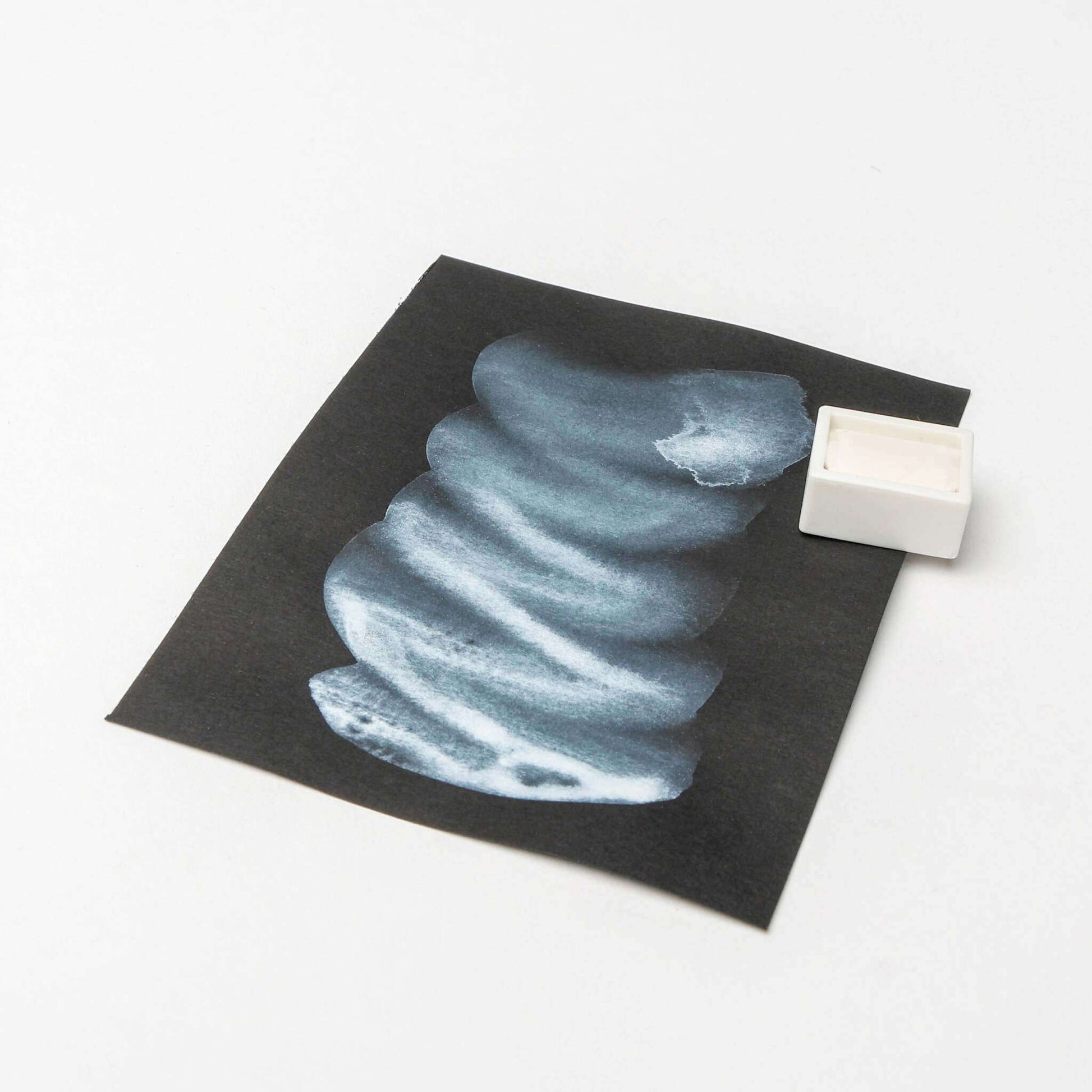 Ein Blatt schwarzes Papier mit blauen Rico-Design-Aquarellfarben neben einem weißen Aquarellfarbkasten.