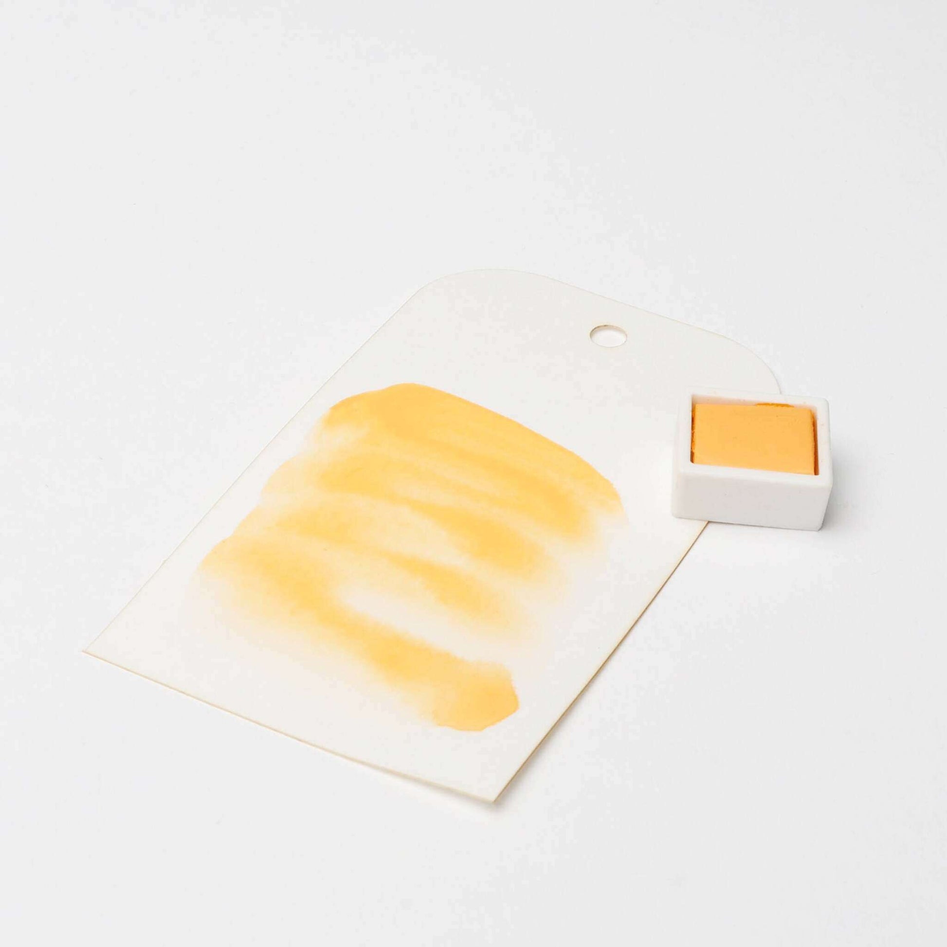 Eine orangefarbene Aquarellfarbe mit kräftigen Strichen liegt neben einer kleinen orangefarbenen Rico Design Künstler Aquarellfarben-Schale auf weißem Hintergrund.