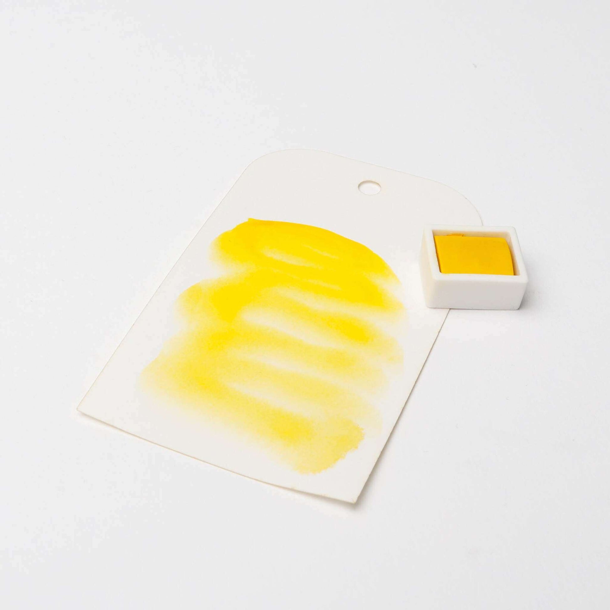 Ein weißes Schild mit gelben Aquarellfarben, auf dem Rico Design Künstler Aquarellfarben anpreist, steht neben einem kleinen quadratischen Topf mit gelber Aquarellfarbe.