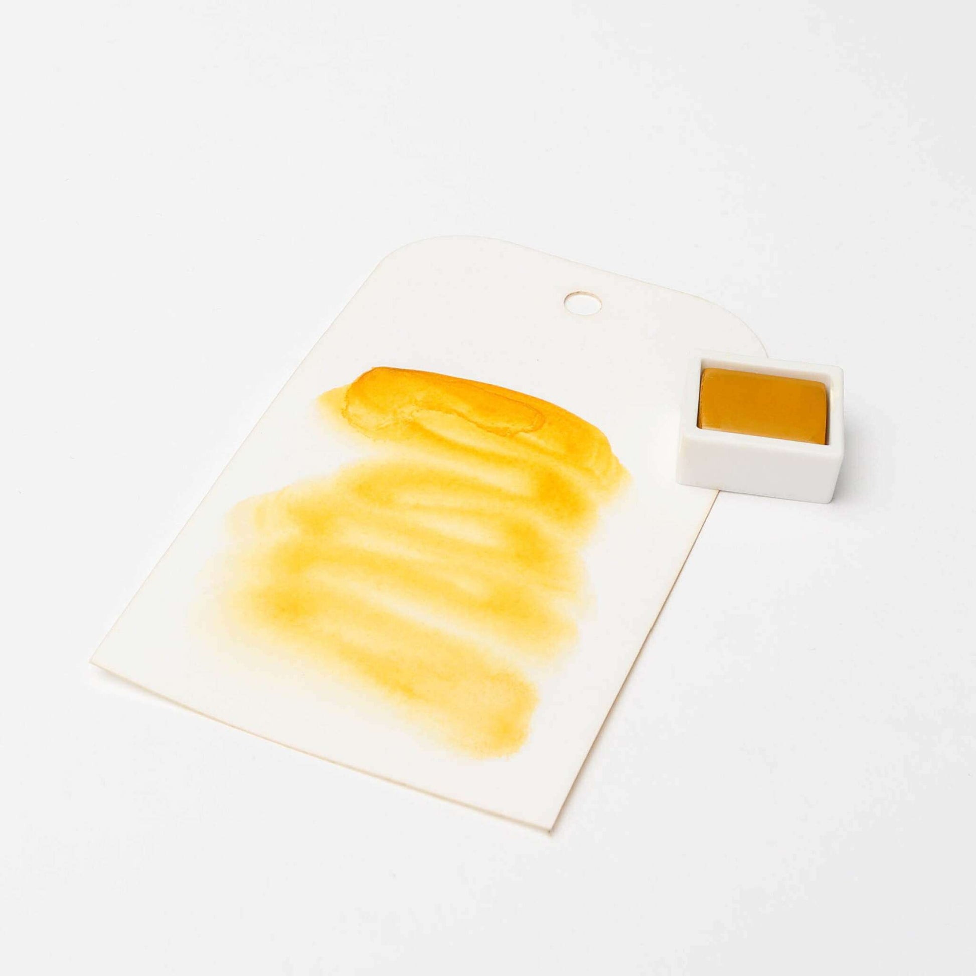 Ein Papier in Form eines Anhängers mit einem gelben Aquarellfarbe-Muster neben einem kleinen Topf mit Rico Design Künstler Aquarellfarben, der die leuchtenden Farbpigmente präsentiert.