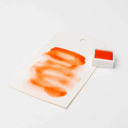 Ein weißer Anhänger mit orangefarbenen Aquarellfarben, einem kleinen Topf mit Rico Design Aquarellfarbe und leuchtenden Farbpigmenten auf einem sauberen weißen Hintergrund.