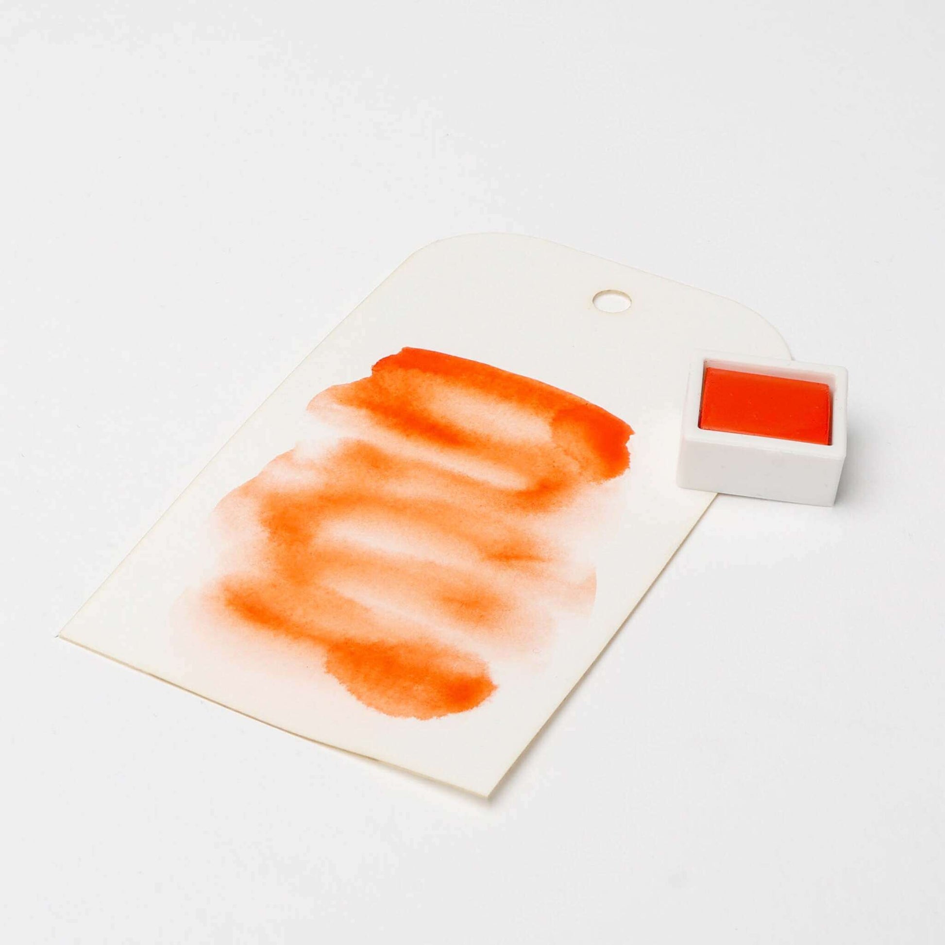 Ein weißer Anhänger mit orangefarbenen Aquarellfarben, einem kleinen Topf mit Rico Design Aquarellfarbe und leuchtenden Farbpigmenten auf einem sauberen weißen Hintergrund.