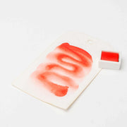 Ein weißes Schild mit einem roten Aquarellmuster, das mit Rico Design hochwertige Künstler Aquarellfarben erstellt wurde, steht neben einem kleinen quadratischen Topf mit roter Farbe auf einer weißen Fläche.