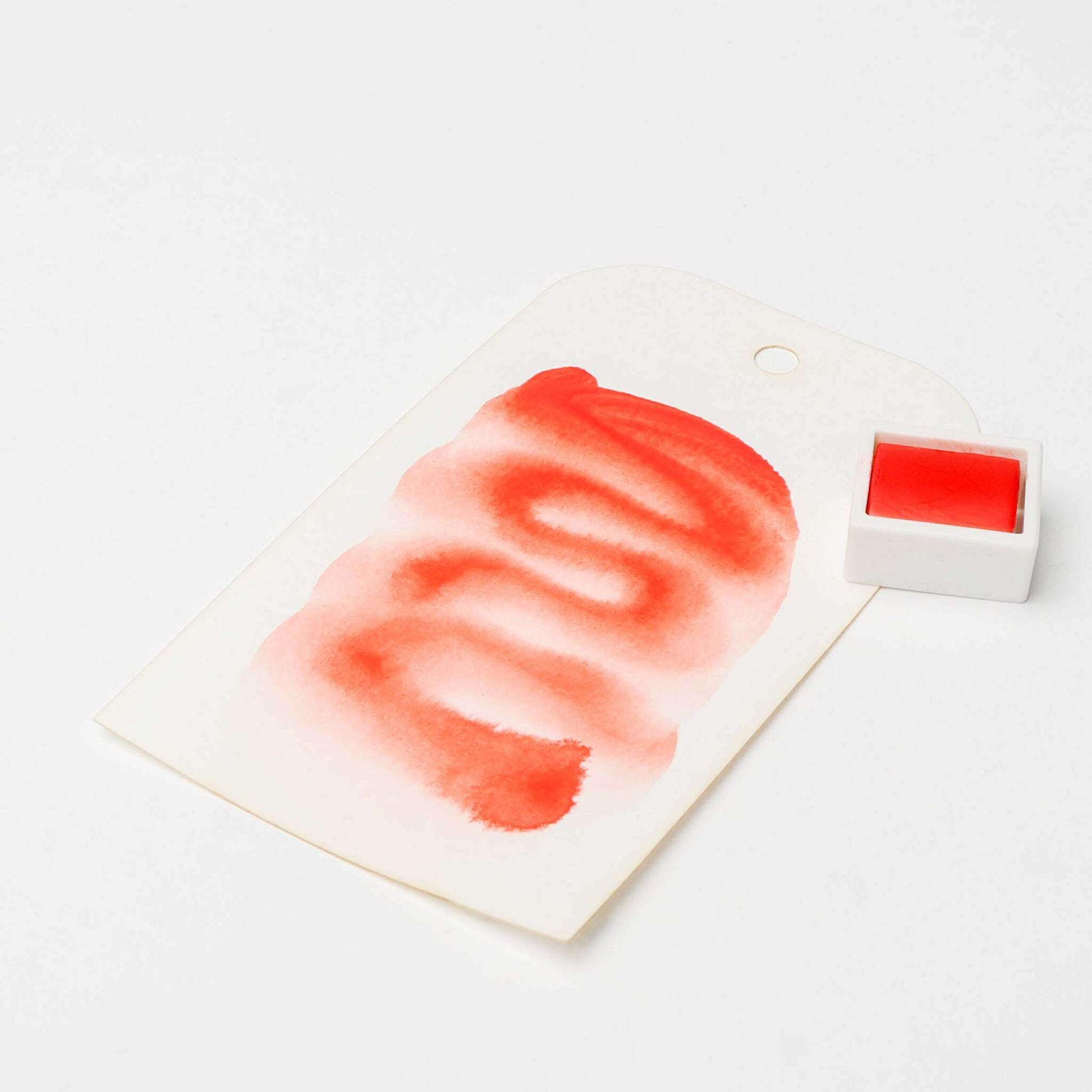 Ein weißes Schild mit einem roten Aquarellmuster, das mit Rico Design hochwertige Künstler Aquarellfarben erstellt wurde, steht neben einem kleinen quadratischen Topf mit roter Farbe auf einer weißen Fläche.