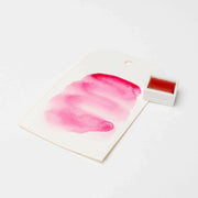 Ein Muster der rosafarbenen Rico Design Künstler Aquarellfarben auf weißem Papier neben einem kleinen passenden Farbtopf zeigt die satten Farbpigmente und die lebendige Qualität.