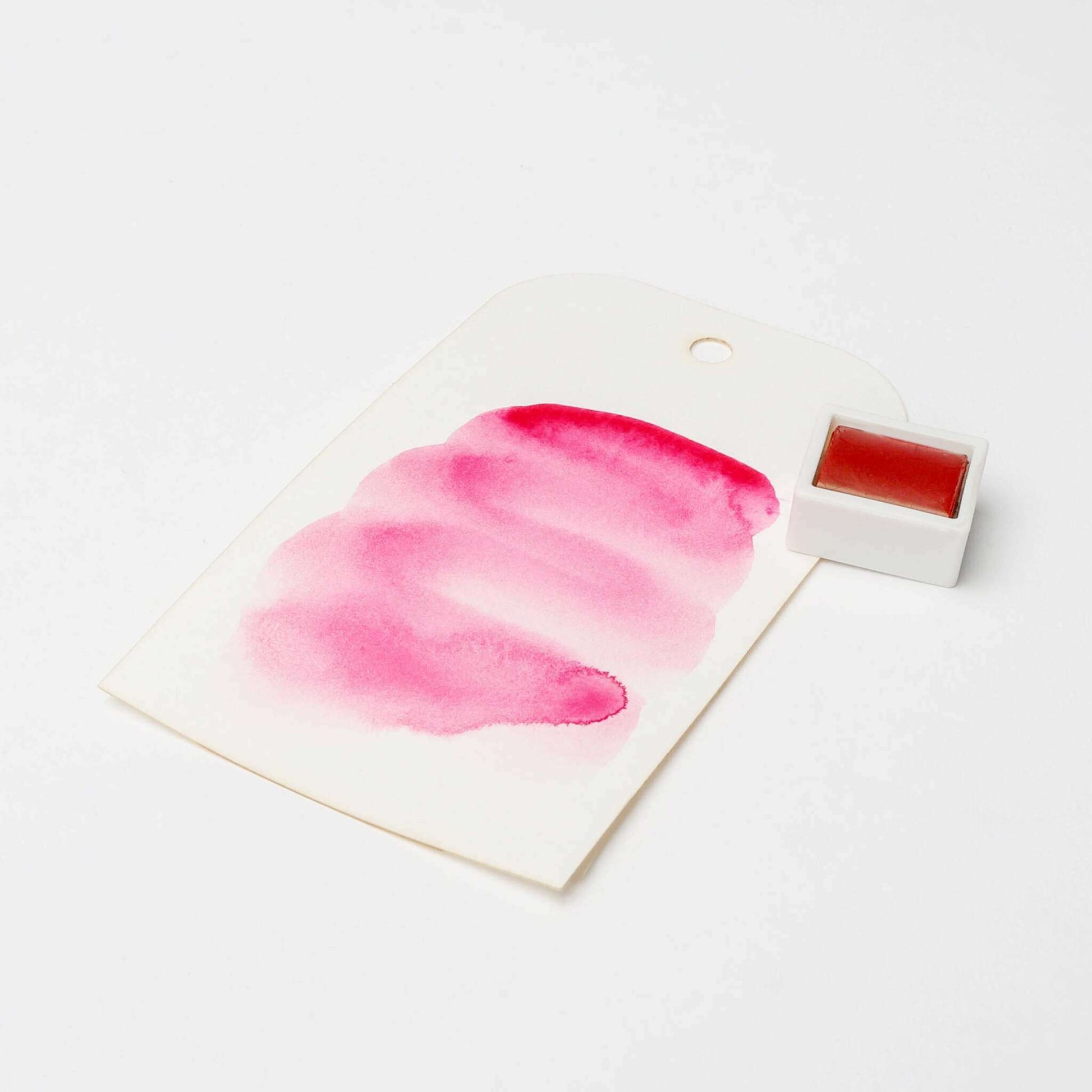 Ein Muster der rosafarbenen Rico Design Künstler Aquarellfarben auf weißem Papier neben einem kleinen passenden Farbtopf zeigt die satten Farbpigmente und die lebendige Qualität.