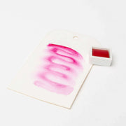 Eine weiße Karte mit rosafarbenen Aquarellfarben steht neben einer kleinen weißen Schale von Rico Design Künstler Aquarellfarben auf weißem Hintergrund.