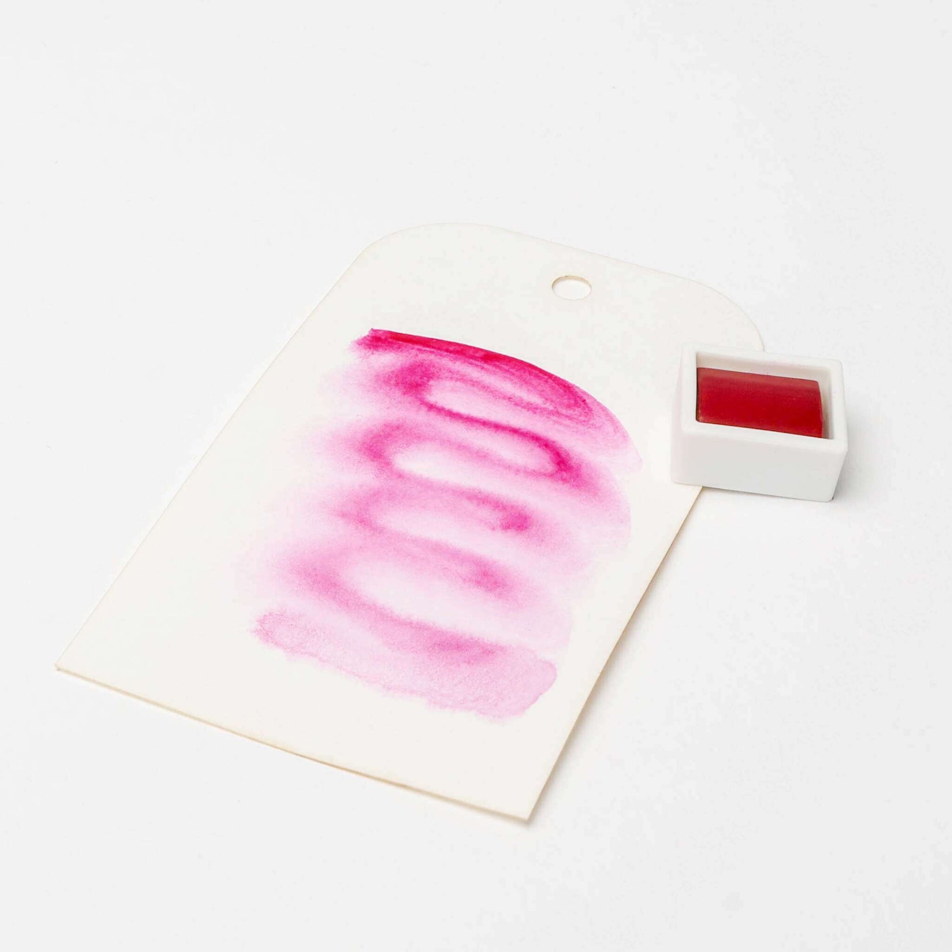 Eine weiße Karte mit rosafarbenen Aquarellfarben steht neben einer kleinen weißen Schale von Rico Design Künstler Aquarellfarben auf weißem Hintergrund.
