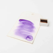 Ein Muster der violetten Rico Design Aquarellfarbe auf einem weißen Anhänger neben einem kleinen Topf mit brauner Farbe, der die reichen Pigmente der Künstler Aquarellfarben zeigt.