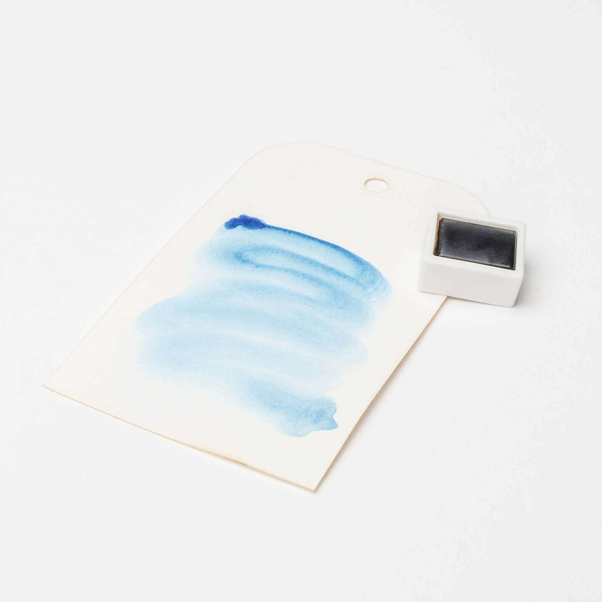 Ein Papieranhänger mit blauer Aquarellfarbe von Rico Design neben einem kleinen schwarzen Farbtopf auf weißem Hintergrund, der die leuchtenden Farbpigmente in Künstler Aquarellfarben hervorhebt.