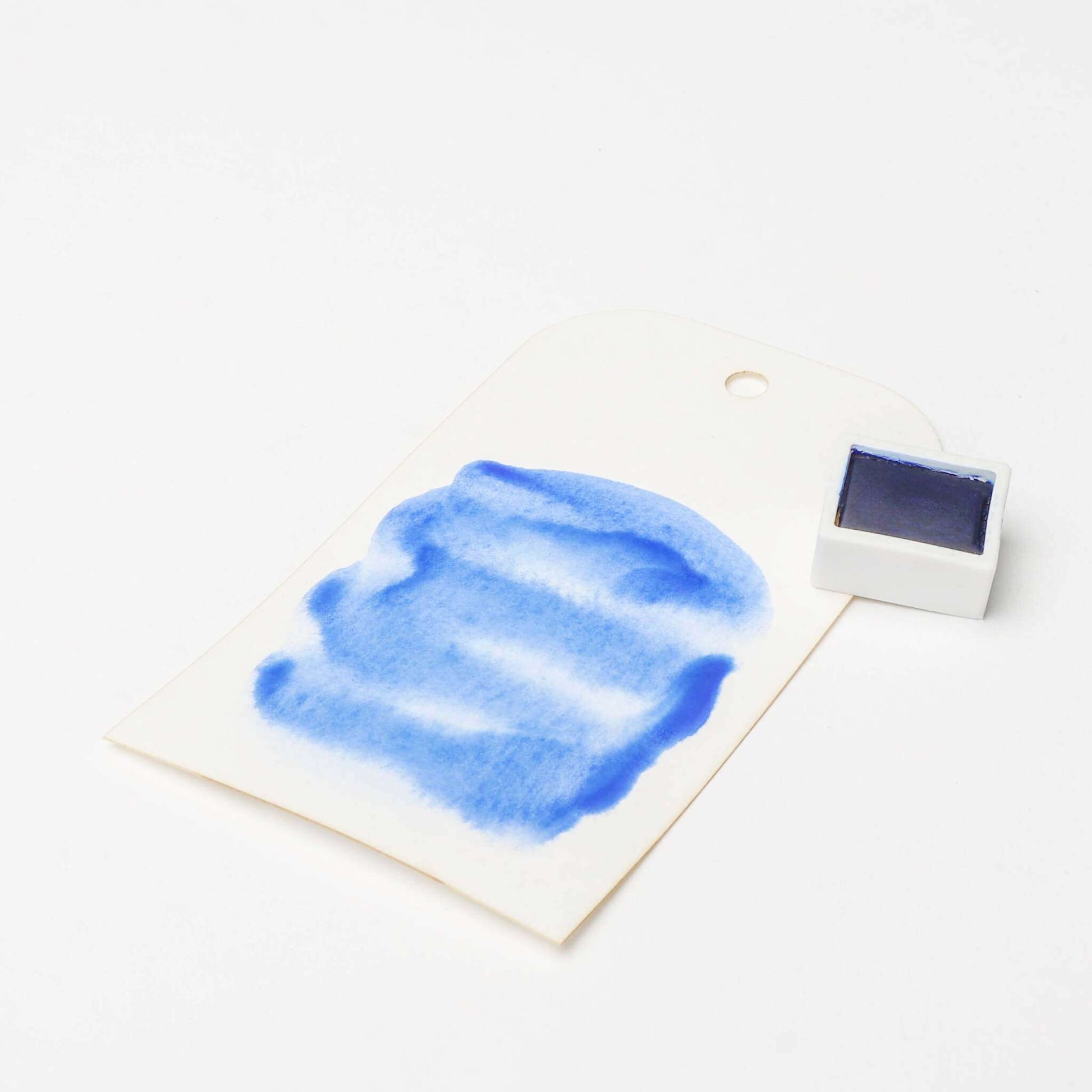 Ein weißer Anhänger mit blauer Aquarellfarbe, der mit Rico Design Künstler Aquarellfarbe gestaltet wurde, steht neben einem kleinen Topf mit Rico Design Aquarellfarben auf weißem Hintergrund.