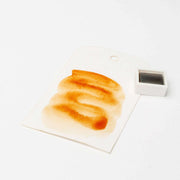 Ein Aquarellmuster in Brauntönen auf einem Anhänger neben einer kleinen Schale mit Rico Design Aquarellfarbe, das die reichen Farbpigmente der Künstler Aquarellfarben zeigt.