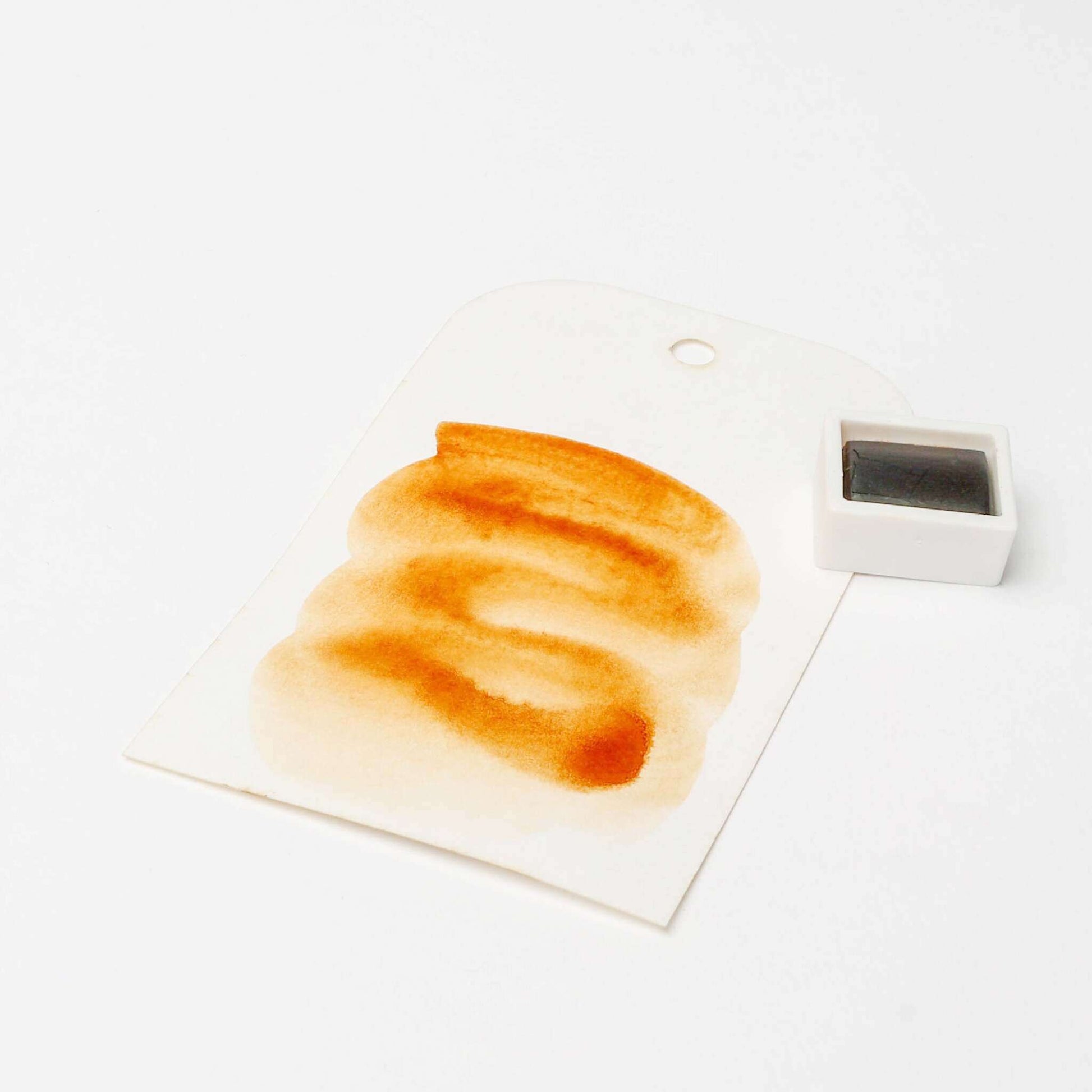 Ein Aquarellmuster in Brauntönen auf einem Anhänger neben einer kleinen Schale mit Rico Design Aquarellfarbe, das die reichen Farbpigmente der Künstler Aquarellfarben zeigt.