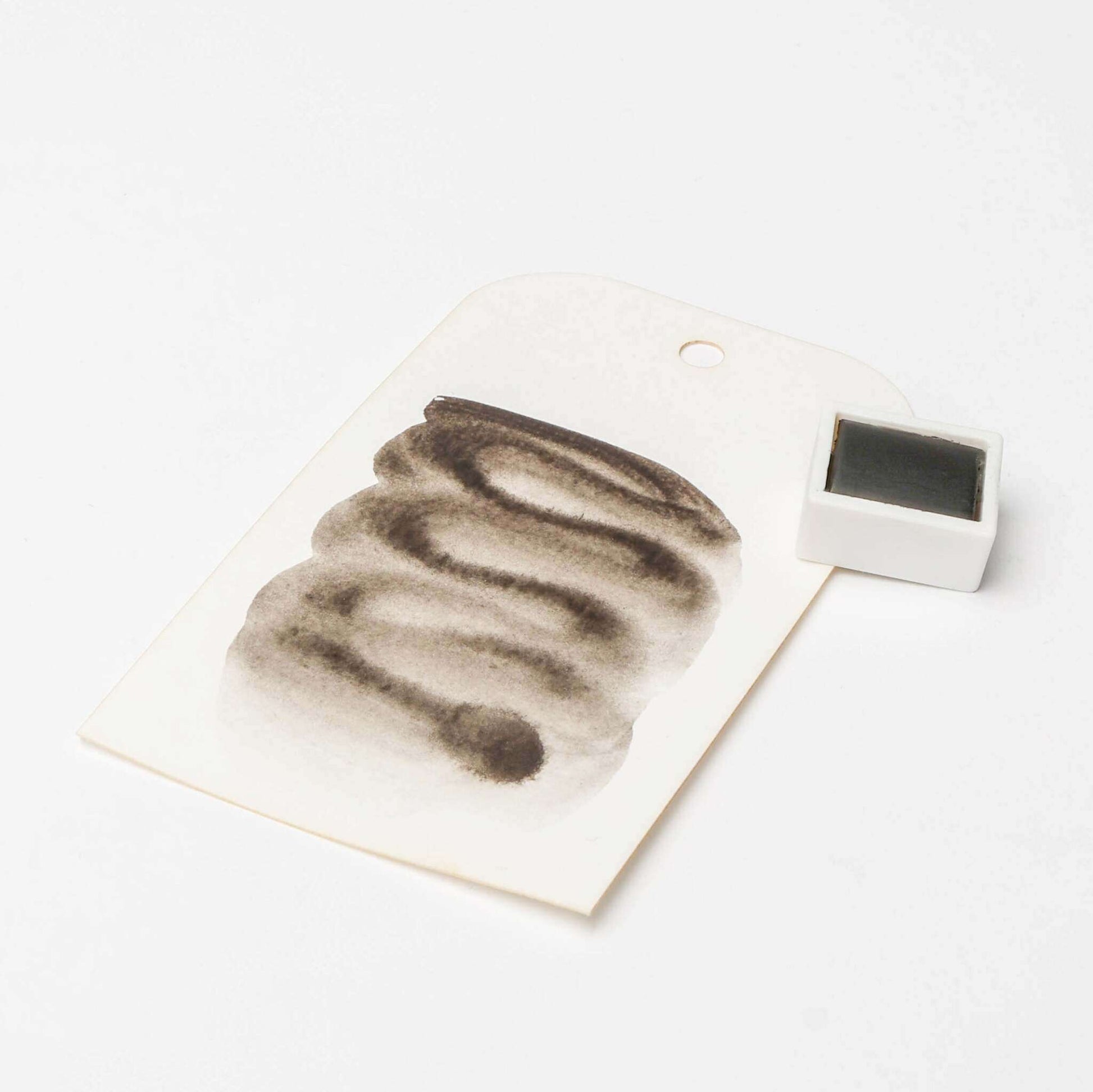 Ein weißer Anhänger mit abstrakter schwarzer Tuschemalerei aus hoch konzentrierten Pigmenten von Rico Design steht neben einem kleinen weißen Stempelkissen auf weißem Hintergrund.