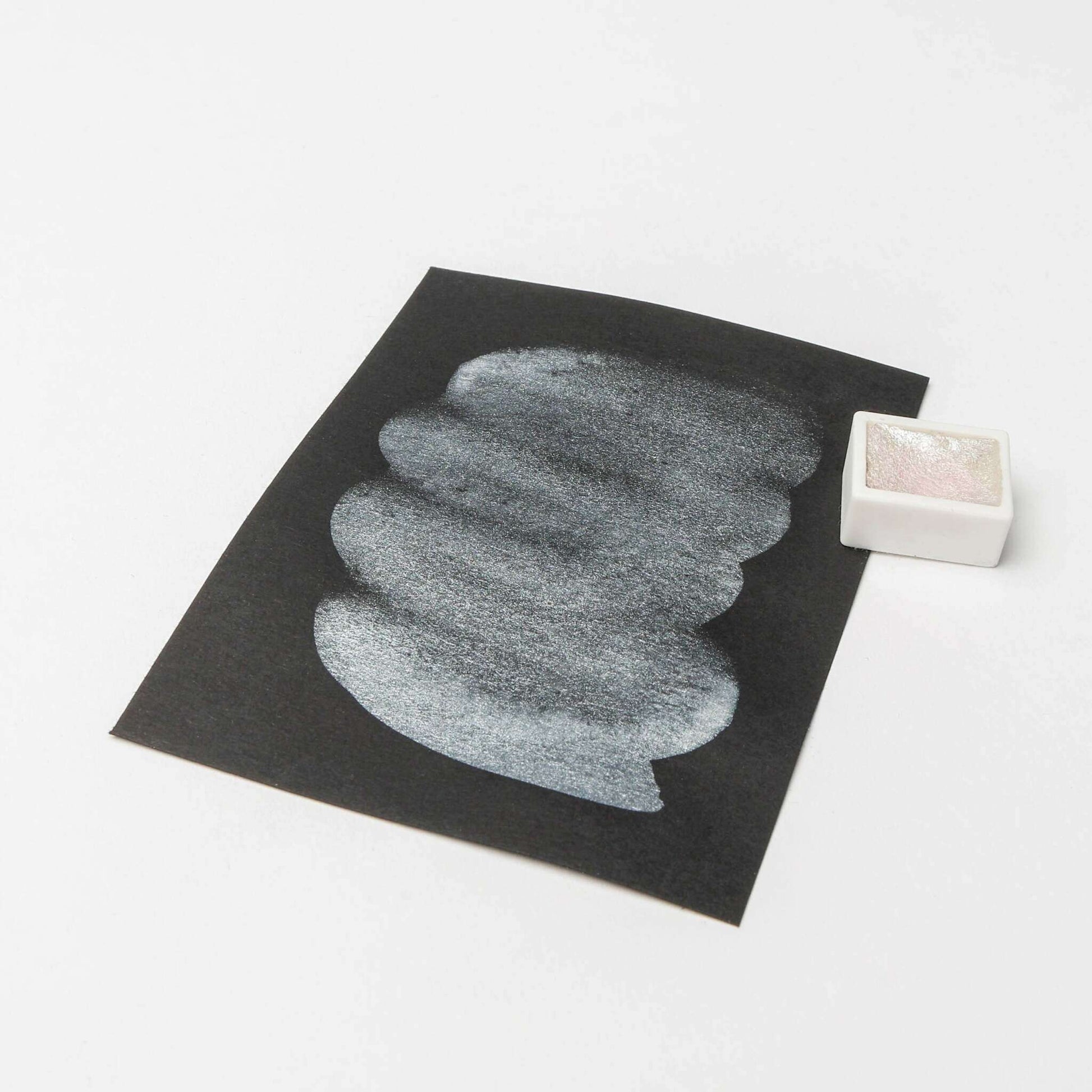 Ein weißer Pastellstrich auf schwarzem Papier liegt neben einem kleinen rechteckigen weißen Behälter von Rico Design, der auf einer weißen Fläche hohe Leuchtkraft und reiche Farbpigmente für lebendige künstlerische Effekte zur Schau stellt.