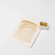 Ein goldenes Aquarellmuster auf einem weißen Anhänger sitzt neben einer kleinen Schale Rico Design Metallic Aquarellfarben und hebt die leuchtenden Metallicfarben vor einem weißen Hintergrund hervor.