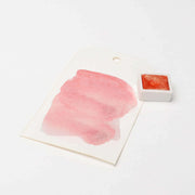 Ein korallenrotes Aquarellmuster von Rico Design mit hoher Deckkraft auf einem weißen Anhänger, abgebildet neben einem kleinen Topf mit hochkonzentrierten Farbpigmenten.