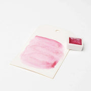 Ein weißes Etikett mit einem rosafarbenen Aquarellmuster steht neben einem kleinen Topf mit rosa schimmernden Metallic-Aquarellfarben von Rico Design, bekannt für seine hohe Deckkraft, auf weißem Hintergrund.