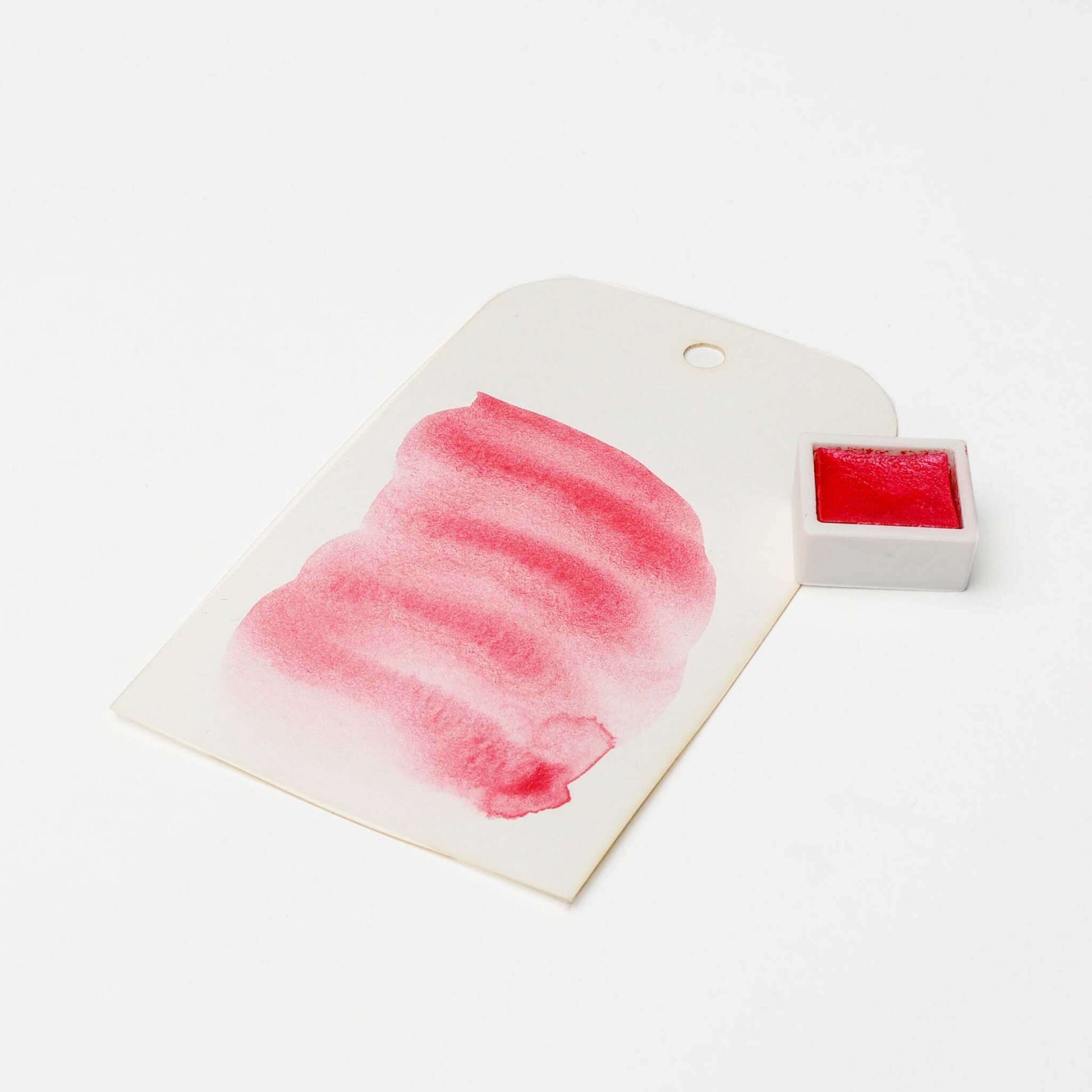 Ein weißes Schild mit einem roten Aquarellmuster steht neben einem kleinen Topf mit hochkonzentrierten Farbpigmenten von Rico Design in sattroter Aquarellfarbe.