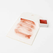 Ein Muster der rot schimmernden Rico Design Metallic Aquarellfarben auf weißem Papier liegt neben einem kleinen Farbtopf und demonstriert seine hohe Leuchtkraft.