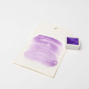 Ein weißes Schild mit einem lila Aquarellmuster und "hoch konzentrierte Farbpigmente" liegt neben einem kleinen Topf mit lila Farbe von Rico Design auf einer weißen Fläche.