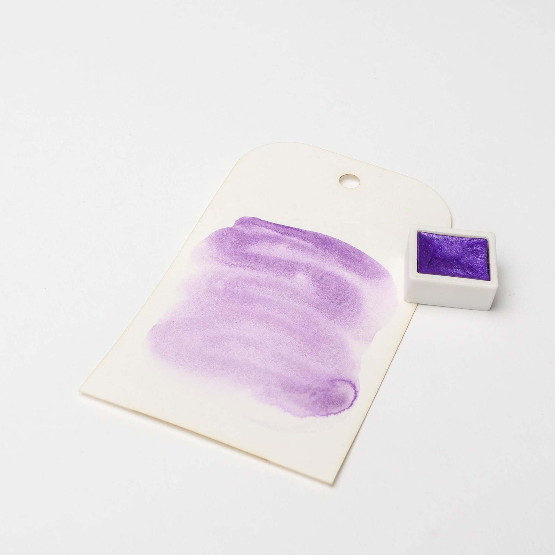 Ein weißes Schild mit einem lila Aquarellmuster und "hoch konzentrierte Farbpigmente" liegt neben einem kleinen Topf mit lila Farbe von Rico Design auf einer weißen Fläche.