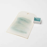 Ein Muster der schimmernden blaugrünen Aquarellfarbe auf Papier liegt neben einer kleinen Schale mit Rico Design Metallic Aquarellfarben, die ihren metallischen Glanz und ihre beeindruckende Lichtechtheit unter Beweis stellt.